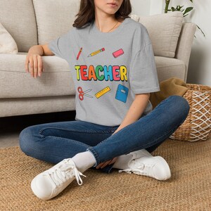 May include: A heather grey t-shirt featuring the word TEACHER in multicoloured block letters. The design includes illustrations of a red crayon, yellow pencil, pink eraser, red scissors, yellow ruler, and a blue notebook surrounding the text.
