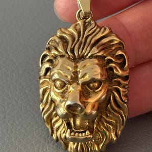 Pode incluir: Um pingente de metal dourado com uma cabeça de leão tridimensional detalhada, com uma juba fluida e a boca aberta mostrando os dentes. O pingente possui uma argola triangular no topo para fixação em uma corrente, com acabamento polido.