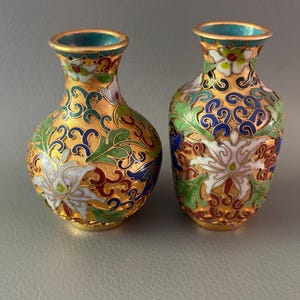 May include: Two small metal vases featuring intricate cloisonné enamel work with a gold-toned base. The surface is decorated with colorful floral patterns in white, green, blue, and red, with a bright turquoise interior lining at the neck.