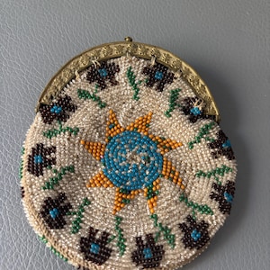 Pode incluir: Uma bolsa de moedas redonda com missangas intrincadas num fundo creme. O design inclui um motivo central de estrela azul e laranja rodeado por padrões florais castanhos escuros e azuis com caules verdes, rematado com uma moldura de latão ornamentada.