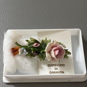 May include: A porcelain floral brooch featuring a sculpted pink rose, green leaves, and a small blue flower, resting on white cotton inside a white plastic box. A small white card with the text CERTIFICATO DI GARANZIA is tucked behind the brooch.