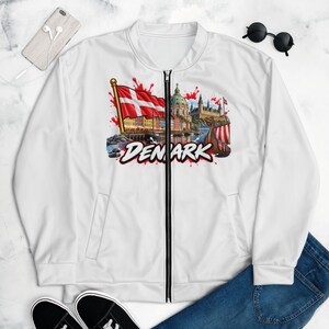 Puede incluir: Una chaqueta bomber blanca con cremallera que presenta un estampado gráfico de la bandera danesa, un paisaje urbano con un edificio con cúpula, un barco vikingo y la palabra DENMARK en letras negras y rojas. Tiene cremallera negra y mangas largas.