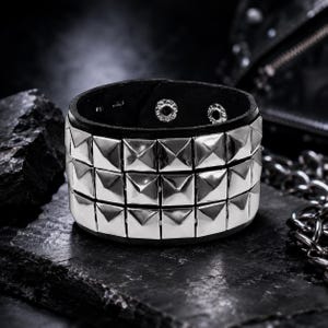 Peut inclure: Un bracelet manchette en cuir noir orné de trois rangées horizontales de clous pyramidaux argentés polis. L'intérieur de la bande présente deux boutons-pression métalliques pour la fermeture, assurant un maintien sûr autour du poignet.