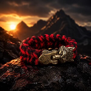 May include: A paracord bracelet featuring a braided pattern of black and red nylon cords. The bracelet is secured with a metallic bronze-toned clasp shaped like a stylized wolf head, showcasing intricate engraved details on the metal surface.