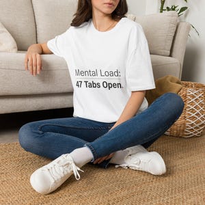 以下が含まれることがあります： 黒い文字で「Mental Load: 47 Tabs Open」とプリントされた白い半袖Tシャツ。このシャツはダークブルーのデニムジーンズと白いスニーカーと組み合わされており、胸の前面に明確なグラフィックデザインが施されています。