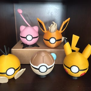 May include: A collection of five spherical figures inspired by Pokemon characters. The set includes a pink Jigglypuff, an orange Eevee, a yellow Electabuzz, a brown-shelled creature, and a yellow Pikachu, each featuring a central white button.