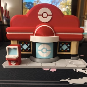 Può includere: Un modello stampato in 3D di un edificio rosso e beige con una porta centrale azzurra dotata di un emblema circolare bianco. La struttura presenta un tetto rosso con un simbolo Poké Ball bianco, due finestre con cornici nere e un piccolo chiosco rosso.