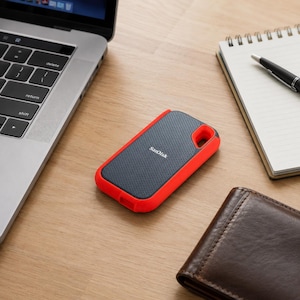 May include: A rectangular SanDisk portable solid state drive featuring a dark gray textured surface with a perforated pattern and a bright orange rubberized protective bumper frame surrounding the edges, positioned on a wooden desk surface.