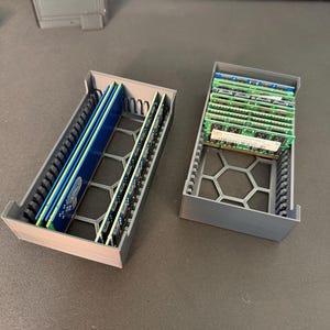 May include: Two 3D-printed gray plastic storage organizers with a hexagonal base pattern. The left organizer holds several long blue RAM sticks, while the right organizer contains multiple smaller green RAM modules arranged in vertical slots.