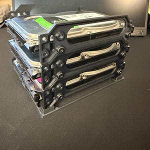 May include: A black 3D-printed plastic storage rack holding three stacked silver and black hard disk drives. The frame features a vertical design with screw-in mounting points on each side, securing the drives in a parallel, tiered arrangement.