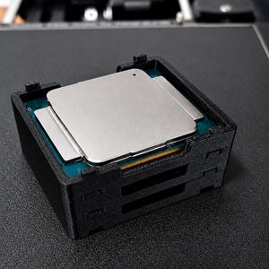 Può includere: Un processore con un dissipatore di calore in metallo argentato e una base in circuito stampato verde, fissato all'interno di una cornice protettiva nera stampata in 3D. Il testo "socket LGA2011" è stampato in bianco nella parte inferiore.