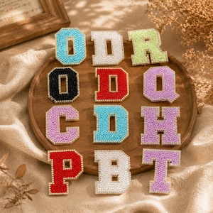 Peut inclure: Une collection de onze écussons en forme de lettres de style universitaire disposés sur une surface en bois. Chaque lettre est recouverte de petits strass étincelants et bordée d'une rangée de minuscules perles blanches. Les lettres incluent O, D, R, O, D, Q, C, D, H, P, B et T.