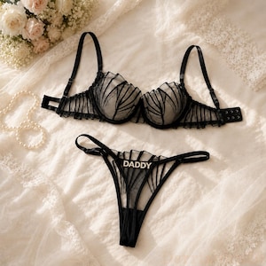 May include: A black lingerie set consisting of a sheer underwire bra and matching thong. Both pieces feature vertical black mesh stripes and ruffled trim. The thong is accented with a silver-toned rhinestone embellishment spelling the word DADDY.