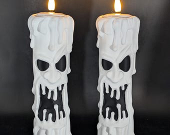 May include: Two white pillar candles with a sculpted, melting wax texture and a carved, menacing face featuring hollow black eyes and a wide, jagged mouth. Each candle is topped with a small, glowing, warm-toned LED flame light.