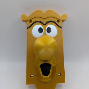 Op de afbeelding: Een gele, rechthoekige deurplaat met een centrale ronde knop, een wijd openstaande mond en grote ogen met zwarte wenkbrauwen. Het object heeft een gladde, gegoten afwerking met zichtbare schroefgaten in de bovenste en onderste hoeken voor montage.