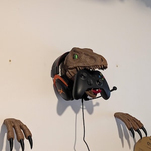 May include: A brown dinosaur head wall mount featuring green eyes and an open mouth holding a black gaming controller. A pair of black and orange over-ear headphones with a microphone is placed on the dinosaur head, which is flanked by two separate brown dinosaur claw wall mounts with black nails.