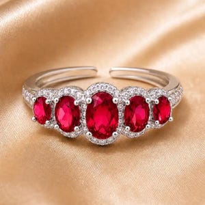 Könnte beinhalten: Ein silberfarbener offener Ring mit fünf oval geschliffenen, leuchtend roten Steinen in einer Reihe. Der mittlere Stein ist am größten und wird von zwei kleineren Steinen auf jeder Seite flankiert. Jeder Stein ist von kleinen weißen Kristallen umgeben.