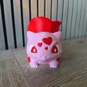 Può includere: Un vaso a forma di creatura rosa chiaro con una grande struttura rossa brillante simile a un bulbo sul dorso. La figura presenta segni rossi a forma di cuore sulla fronte e sui lati, con occhi rossi e artigli bianchi sui piedi come contenitore.