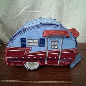 May include: A fabric applique shaped like a vintage camper trailer. The design features a light blue upper section with dark blue shutters and a red lower section with white circular accents. It includes a grey wheel and a red awning over the door.