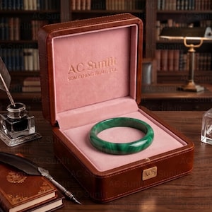 Puede incluir: Un brazalete circular de jade verde liso descansa dentro de una caja de cuero marrón abierta con un interior de terciopelo rosa suave. La tapa presenta el texto en relieve AC Sunlit SOMETHING BEAUTIFUL y una placa dorada con las siglas ACS.