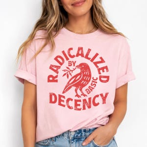 May include: A light pink short-sleeved t-shirt featuring a red graphic of a bird holding a branch. The text RADICALIZED BY DECENCY is printed in a circular, distressed red font around the bird illustration, centred on the chest of the shirt.