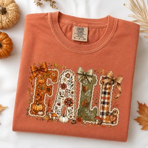 May include: A burnt orange short-sleeve t-shirt featuring the word FALL in large decorative letters. Each letter is adorned with autumn-themed illustrations including pumpkins, mushrooms, leaves, and flowers, topped with bows. The neck label reads COMFORT COLORS.