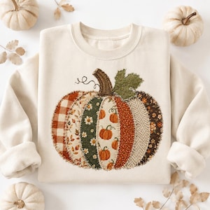 Op de afbeelding: Een crèmekleurige sweater met ronde hals en een grote, centrale pompoenafbeelding. De pompoen is samengesteld uit diverse stofachtige patches in tinten oranje, groen, bruin en wit, met patronen zoals ruiten, bloemen en visgraat.