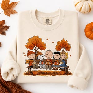 May include: A cream-colored crewneck sweatshirt featuring a graphic of the Peanuts characters sitting on a wooden bench under autumn trees, each holding a mug. The characters include Charlie Brown, Snoopy, Linus, Lucy, and Sally.