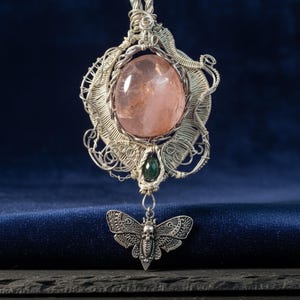 Op de afbeelding: Een zilveren, met draad omwikkelde hanger met een grote, ovale, doorschijnende perzikkleurige steen en een kleine, donkergroene druppelvormige steen. Een zilverkleurige mot-bedel met schedeldetail hangt onderaan de met draad omwikkelde zetting.