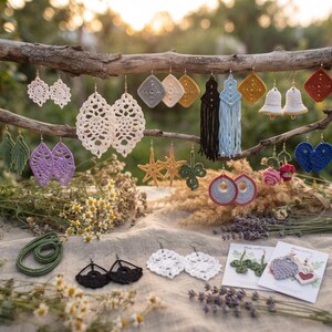 May include: A collection of handmade crochet earrings in various shapes and colors, including leaf, star, flower, and geometric designs. The earrings are displayed hanging from a wooden branch and laid out on a light-colored fabric surface.