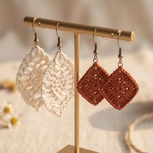 May include: Two pairs of crochet earrings hanging on a gold-toned metal T-bar stand. One pair features cream-colored leaf-shaped motifs, while the other pair consists of rust-colored square motifs. Each earring is attached to a metal hook finding.