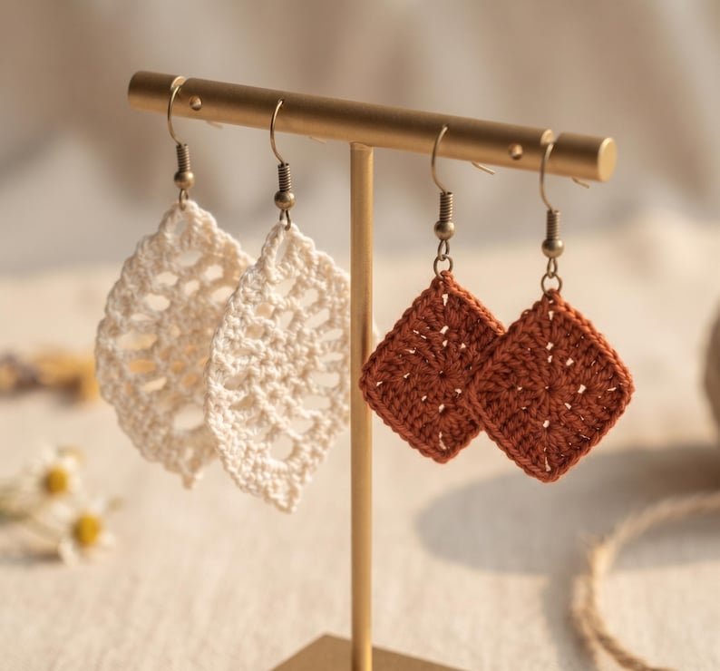May include: Two pairs of crochet earrings hanging on a gold-toned metal T-bar stand. One pair features cream-colored leaf-shaped motifs, while the other pair consists of rust-colored square motifs. Each earring is attached to a metal hook finding.