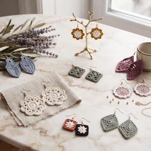 May include: A collection of various crocheted earrings in diverse shapes and colors, including white floral, blue leaf-shaped, green square, and pink circular designs, displayed alongside a gold-toned jewelry stand and a small gold chain.