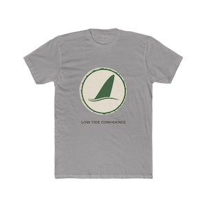 May include: A grey short-sleeve t-shirt featuring a circular graphic on the chest. The graphic contains a dark green shark fin icon over a cream background with a distressed border. Below the circle, the text reads LOW TIDE CONFIDENCE.