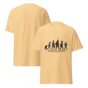 Op de afbeelding: Een lichtgeel t-shirt met korte mouwen en een zwarte grafische print van de menselijke evolutie, van een primaat tot een moderne soldaat. De tekst onder de figuren luidt THE PEAK OF EVOLUTION, met een pijl die naar de centrale figuur wijst.