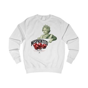 Puede incluir: Una sudadera blanca de manga larga con un gráfico de un monarca señalando dados rojos marcados con símbolos de divisas. El texto dice MISPRINTED DEPT y BUILT DIFFERENT. El diseño incluye una corona, un collar y una prenda de color verde.