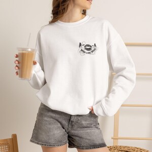 Op de afbeelding: Een wit sweatshirt met lange mouwen en een ronde hals, voorzien van een kleine afbeelding op de linkerborst. De afbeelding toont een paar lippen omringd door decoratieve krullen en de tekst WAH LUH WAH in een zwart, gotisch lettertype.