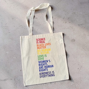 Può includere: Una borsa tote in tela color crema con due manici, caratterizzata da un testo multicolore che recita: SCIENCE IS REAL, BLACK LIVES MATTER, NO HUMAN IS ILLEGAL, LOVE IS LOVE, WOMEN'S RIGHTS ARE HUMAN RIGHTS e KINDNESS IS EVERYTHING.