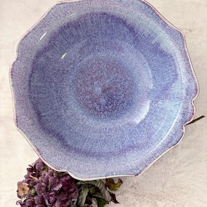 May include: A ceramic bowl with a scalloped edge and a mottled glaze featuring shades of light blue, lavender, and deep purple. The interior surface displays a speckled texture that transitions from a dark center to a lighter rim.