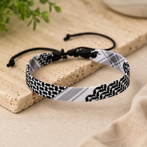 May include: A black and white woven fabric bracelet featuring a mix of geometric patterns including houndstooth, diagonal stripes, and a wavy chevron design. The bracelet has a black cord closure with adjustable sliding knots for a secure fit.