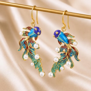 May include: A pair of gold-tone fish-shaped drop earrings featuring vibrant blue, purple, and red enamel detailing. Each earring is accented with multiple small white spherical pearls along the flowing tail fins and hangs from a simple hook wire.