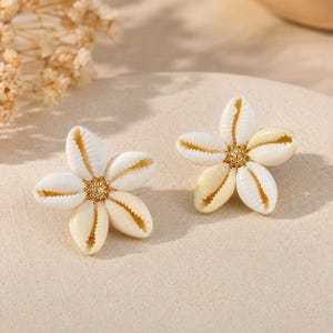 May include: A pair of stud earrings shaped like flowers, each constructed from five white cowrie shells arranged around a central gold-toned circular component with a textured surface. The shells are smooth, off-white, and oval-shaped.