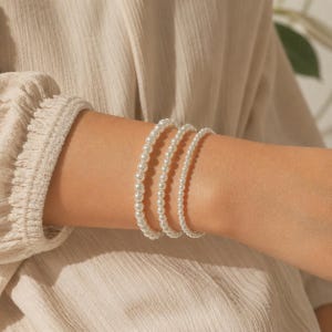 May include: Three stacked bracelets made of round white pearls of varying sizes. The bracelets feature a uniform, smooth, and glossy finish, arranged in a sequence from the largest beads to the smallest beads on the wrist, creating a layered look.