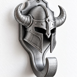 May include: A silver-toned wall hook featuring a detailed Viking helmet design with two prominent curved horns. The helmet is mounted on a shield-shaped backplate with a visible screw at the top, and a curved hook extends from the bottom section.