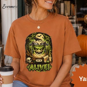 May include: A burnt orange t-shirt featuring a graphic print of a green monster in a glass jar with the text IT'S ALIVE! The design includes a skull, a raven, and lightning bolts in shades of green, yellow, and black against a dark background.