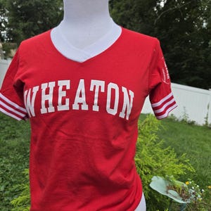 May include: A red short-sleeve V-neck t-shirt with white trim on the collar and sleeves. The word WHEATON is printed in large white block letters across the chest. A small white graphic of a baseball is visible on the left sleeve of the shirt.