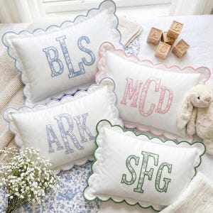 May include: Four rectangular white pillows with scalloped edges, each featuring a three-letter monogram embroidered in a floral pattern. The monograms are blue, pink, purple, and green. Wooden alphabet blocks and a plush toy are nearby.