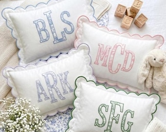May include: Four rectangular white pillows with scalloped edges, each featuring a three-letter monogram embroidered in a floral pattern. The monograms are blue, pink, purple, and green. Wooden alphabet blocks and a plush toy are nearby.