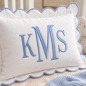 Op de afbeelding: Een rechthoekig wit kussen met een geschulpte rand afgewerkt met lichtblauw garen. Het midden van het kussen toont een groot, geborduurd monogram in lichtblauw garen met de letters KMS in een klassieke schreeflettertypestijl.