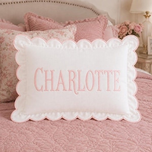 Peut inclure: Un coussin rectangulaire blanc avec un bord festonné et une bordure rose. Le centre du coussin est brodé du nom CHARLOTTE dans une police serif rose. Le coussin présente une finition en tissu texturé et une bordure décorative.
